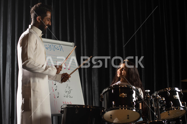 تقديم البرامج والفنون الموسيقية والفن المسرحي، الاشكال الايقاعية الموسيقية، رجل عربي خليجي سعودي يقوم بتعليم الطفلة على آلة الطبل، جلسات تدريب في الموسيقى، ممارسة هواية الطبل باستخدام آلة الطبل الالكترونية، مواهب فنية موسيقية، انشطة ترفيهية