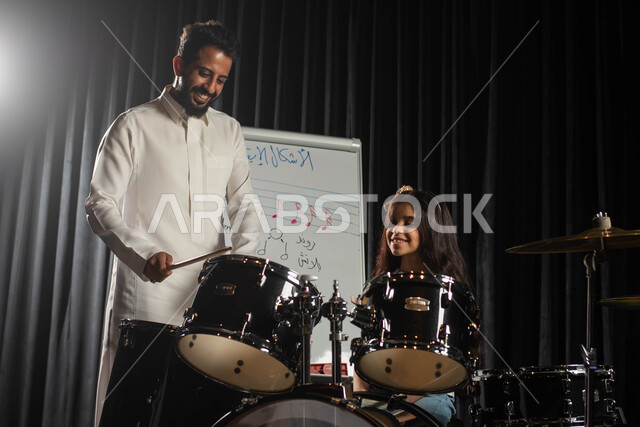 ممارسة هواية الطبل باستخدام آلة الطبل الالكترونية، الاشكال الايقاعية الموسيقية، رجل عربي خليجي سعودي يقوم بتعليم الطفلة على آلة الطبل، جلسات تدريب في الموسيقى، تقديم البرامج والفنون الموسيقية والفن المسرحي، مواهب فنية موسيقية، انشطة ترفيهية