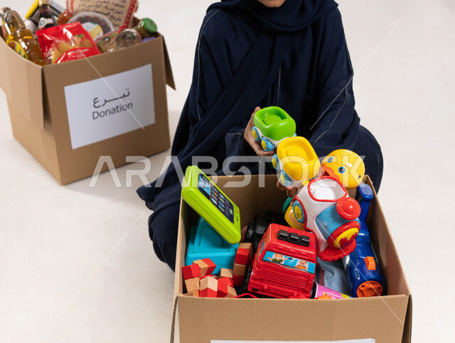 A close-up of a Saudi Arabian Gulf woman preparing a donation box full of children's toys, providing toys for needy children, donating to the food basket project, charitable humanitarian initiatives and donations, Islamic associations in Saudi Arabia, whi