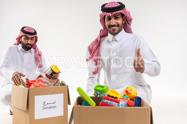 بورتريه لرجل يرفع اصبع السبابة بايماءات تدل على الاعجاب، القبول والموافة، رجلان عربيان خليجيان سعوديان، صندوق مليئ بالعاب الاطفال، التبرع لمشروع السلة الغذائية، مبادرات وتبرعات انسانية خيرية، جمعيات اسلامية في السعودية، شهر رمضان، خلفية بيضاء