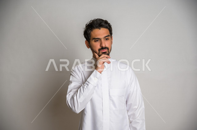 بورتريه لرجل عربي سعودي خليجي باللباس التقليدي , يقوم بإيماءات وجه تدل على التفكير و التركيز ، خلفية بيضاء .