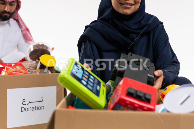 صورة مقربة لامرأة ورجل عربيان خليجيان سعوديان، صندوق تبرعات مليء بالعاب الاطفال، التبرع لمشروع السلة الغذائية، مبادرات وتبرعات انسانية خيرية، جمعيات اسلامية في السعودية، شهر رمضان، خلفية بيضاء