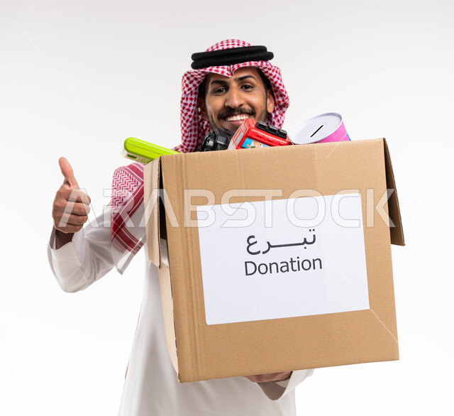 بورتريه لرجل عربي خليجي سعودي يحمل صندوق تبرعات مليء بالعاب الاطفال ويرفع اصبع السبابة لاعلى، ايماءات وجه ويدين تدل على الاعجاب والقبول والموافقة، مبادرات وتبرعات انسانية خيرية، جمعيات اسلامية في السعودية، خلفيه بيضاء