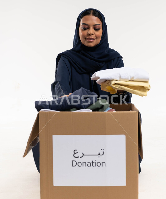 Portrait of a Saudi Arab Gulf woman preparing a donation box for surplus clothes, gifting Eid clothes to the needy, collecting and distributing charitable donations, charities and humanitarian initiatives, serving the needy Saudi society, white background