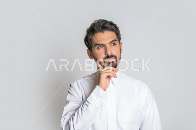 بورتريه لرجل عربي سعودي خليجي باللباس التقليدي , يقوم بإيماءات وجه تدل على التفكير و التركيز ، خلفية بيضاء .