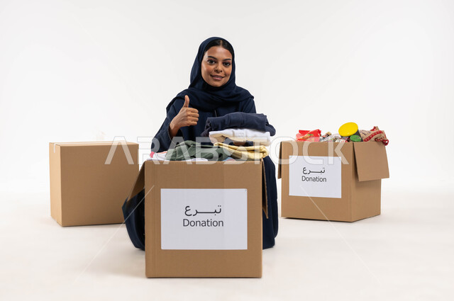 ترتيب ملابس وهدايا العيد السعيد، بورتريه لامرأة عربية خليجية سعودية ترفع اصبع السبابة لاعلى بايماءات تدل على القبول والموافقة، جمع وتوزيع صناديق التبرعات للمحتاجين، مبادرات وتبرعات انسانية خيرية، جمعيات اسلامية في السعودية، خلفية بيضاء