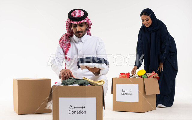 بورتريه لامرأة ورجل عربيان خليجيان سعوديان يتشاركان في جمع وتوزيع صناديق التبرعات للمحتاجين، ترتيب ملابس وهدايا العيد السعيد، مبادرات وتبرعات انسانية خيرية، جمعيات اسلامية في السعودية، خلفيه بيضاء