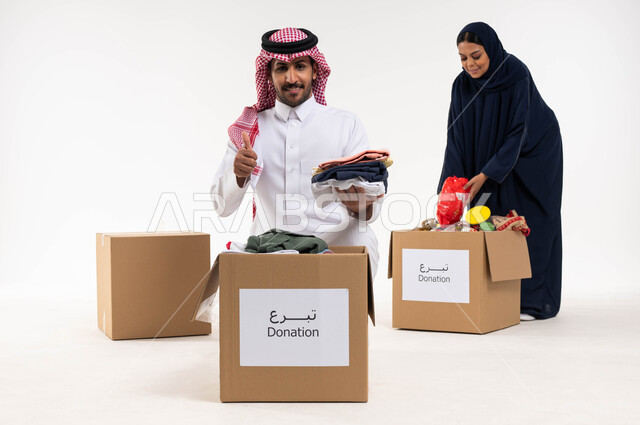 بورتريه لرجل وامرأة عربيان خليجيان سعوديان يقومان بجمع وتوزيع صناديق التبرعات للمحتاجين، رجل بايماءات تدل على القبول والموافقة، مبادرات وتبرعات انسانية خيرية، ترتيب ملابس وهدايا العيد السعيد، جمعيات اسلامية في السعودية، خلفيه بيضاء