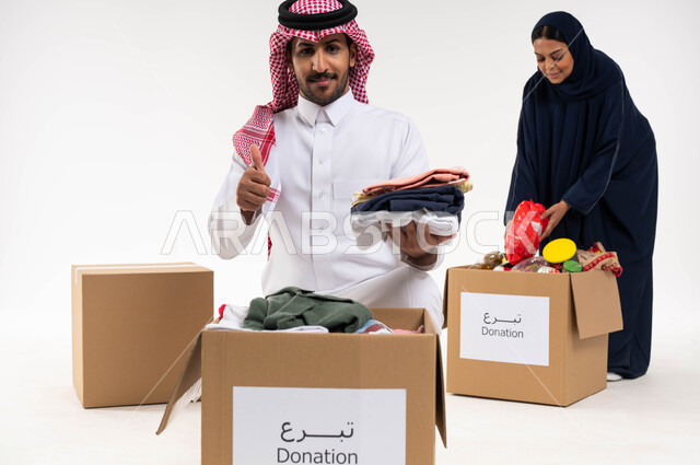 بورتريه لرجل بايماءات تدل على القبول والموافقة، رجل وامرأة عربيان خليجيان سعوديان يقومان بجمع وتوزيع صناديق التبرعات للمحتاجين، مبادرات وتبرعات انسانية خيرية، ترتيب ملابس وهدايا العيد السعيد، جمعيات اسلامية في السعودية، خلفيه بيضاء