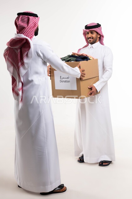 بورتريه لرجلان عربيان خليجيان يتشاركان في تجهيز صندوق تبرعات الملابس الفائضة، اهداء ملابس العيد للمحتاجين، جمع وتوزيع التبرعات الخيرية، جمعيات خيرية ومبادرات انسانية، خدمة المجتمع السعودي المتعفف، صورة الجسم بالطول الكامل، خلفية بيضاء