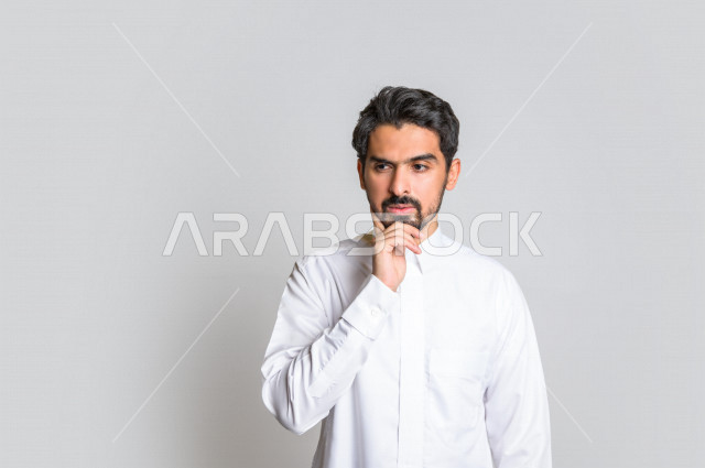 بورتريه لرجل عربي سعودي خليجي باللباس التقليدي , يقوم بإيماءات وجه تدل على التفكير و التركيز ، خلفية بيضاء .
