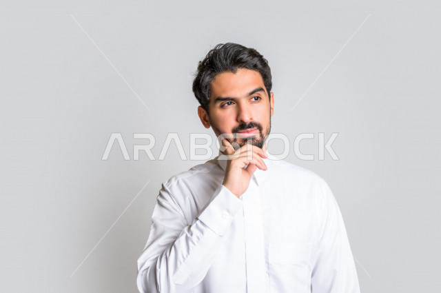 بورتريه لرجل عربي سعودي خليجي باللباس التقليدي , يقوم بإيماءات وجه تدل على التفكير و التركيز ، خلفية بيضاء .