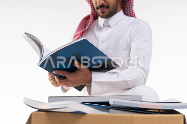 توفير الكتب التعليميه للمحتاجين ، بورتريه مقرب لرجل خليجي  عربي سعودي يرتدي الزي التقليدي  يقوم بتصفح واختيار كتاب من صندوق التبرعات ، التبرع بالكتب الفائضة ،  تبرعات  ومبادرات انسانية ،اعمال خيرية اسلاميه  في السعودية ، خلفية بيضاء.