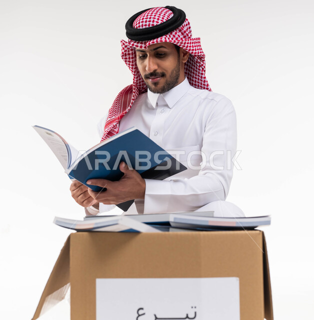 تبرعات  ومبادرات انسانية ، بورتريه لرجل  خليجي عربي سعودي يرتدي الزي التقليدي  يقوم بتصفح واختيار كتاب من صندوق التبرعات ، التبرع بالكتب الفائضة ، توفير الكتب التعليميه للمحتاجين ،  اعمال خيرية اسلاميه  في السعودية ، خلفية بيضاء.
