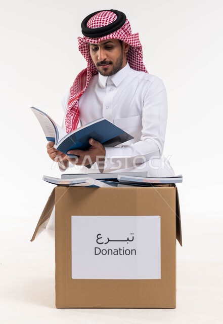 التبرع بالكتب الفائضة  ، بورتريه لرجل  خليجي عربي سعودي يرتدي الزي التقليدي  يقوم بتصفح واختيار كتاب من صندوق التبرعات ، تبرعات  ومبادرات انسانية ، توفير الكتب التعليميه للمحتاجين ،  اعمال خيرية اسلاميه  في السعودية ، خلفية بيضاء.
