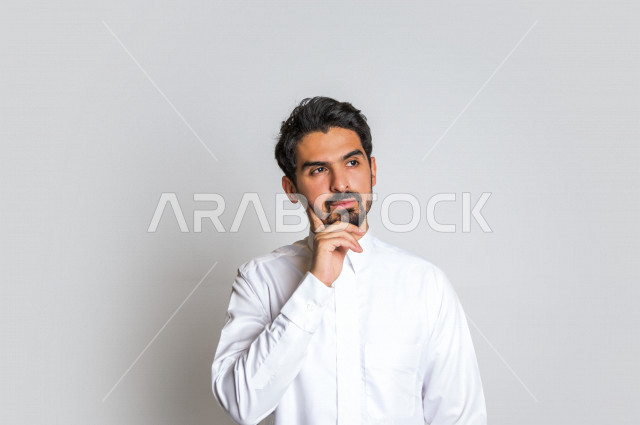 بورتريه لرجل عربي سعودي خليجي باللباس التقليدي , يقوم بإيماءات وجه تدل على التفكير و التركيز ، خلفية بيضاء .