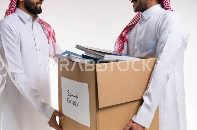 بورتريه لرجلان عربيان خليجيان سعوديان يرتديان الزي السعودي التقليدي يقومان بحمل  صندوق مليئ بالكتب المفيدة ، اعادة التدوير واستخدام الكتب ، التبرع بالكتب الفائضة ، اعمال خيرية اسلاميه  في السعودية  ، خلفية بيضاء.