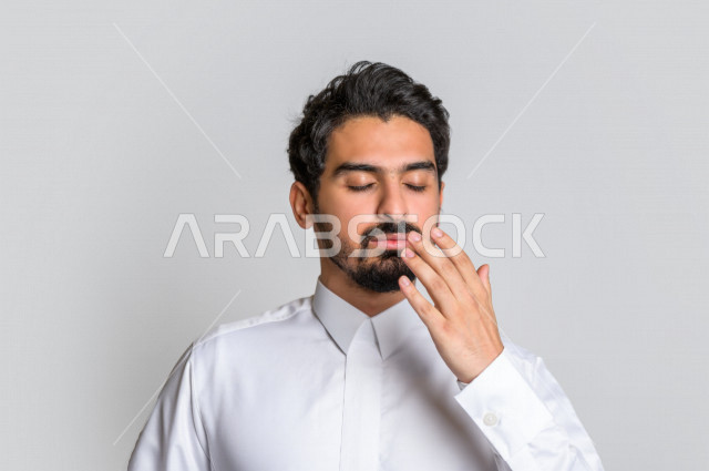 بورتريه لرجل عربي سعودي خليجي باللباس التقليدي السعودي , إيماءات وجه ويدين تدل على النعاس , الإحساس بالتعب والتثاؤب , الحاجة للنوم والراحة , الكسل والخمول ,خلفية بيضاء .