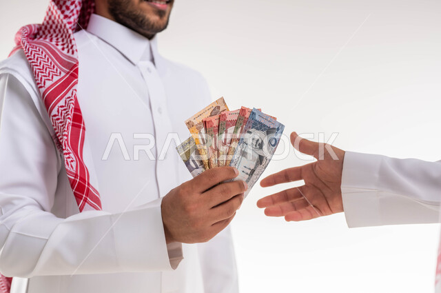 بورتريه مقرب لرجل سعودي عربي  خليجي يرتدي الزي التقليدي يقوم بتقديم مجموعة من النقود الورقية ، اخراج زكاة المال في شهر رمضان ، جمعيات خيرية اسلاميه  في السعودية ، خلفية بيضاء.