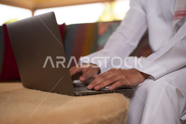 عقد إجتماع عمل عبر الإنترنت ، شاب عربي خليجي سعودي يجلس في المنزل يستخدم الكومبيوتر ، إستخدام جهاز الحاسوب المحمول، تصفح الإنترنت، جلسة عمل، العمل عن بعد ، التسويق الإلكتروني، مهن ووظائف