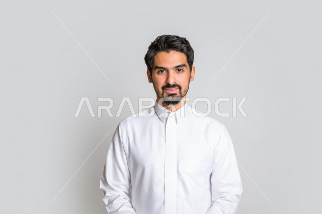 بورتريه لرجل عربي سعودي خليجي باللباس التقليدي , يقوم بالإبتسام أمام الكاميرا لإلتقاط صورة تذكارية , إيماءات وجه تدل على السعادة ، خلفية بيضاء . .