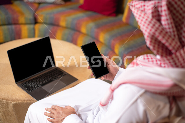 استخدام جهاز الموبايل ، شاب عربي خليجي سعودي مبتسم يرتدي الزي التقليدي السعودي بإيماءات و تعابير وجه تدل على السعادة يتكلم على الهاتف المحمول ، استخدام الحاسوب المحمول ، تواصل ودردشة