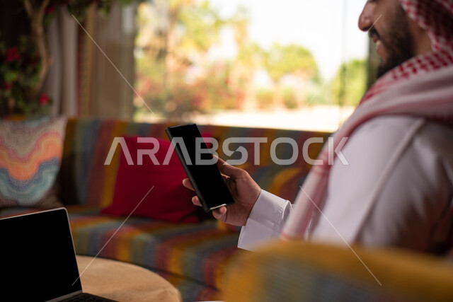 إيماءات و تعابير وجه تدل على السعادة  ، شاب عربي خليجي سعودي مبتسم يرتدي الزي التقليدي السعودي يتكلم على جهاز  الموبايل ، استخدام الهاتف المحمول ، تواصل ودردشة