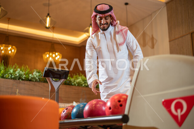 ايماءات وجه تدل على الفرح والسرور ، قضاء أوقات ممتعة في ممارسة لعبة البولينج ، أنشطة رياضية ترفيهية ، رجل عربي خليجي سعودي يرتدي الزي السعودي التقليدي يمارس لعبة البولينج ( الكردحه ) ،في أماكن ترفيهية ، مضمار البولينج ، ملعب البولينج