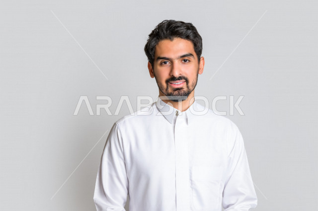 بورتريه لرجل عربي سعودي خليجي باللباس التقليدي , يقوم بالإبتسام أمام الكاميرا لإلتقاط صورة تذكارية , إيماءات وجه تدل على السعادة ، خلفية بيضاء . .