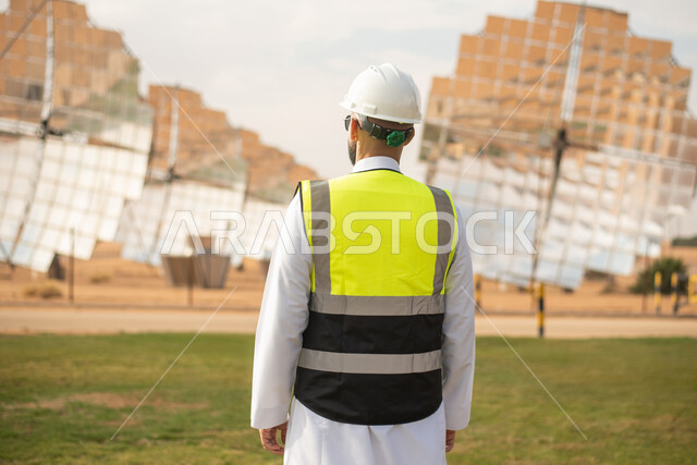 صورة من الخلف لمهندس عربي خليجي سعودي، يرتدي الخوذة وسترة الحماية، ، التدقيق والإشراف على العمل، مفهوم هندسة الطاقة، مهن ووظائف سعودية، توليد الكهرباء عبر محطات الطاقة الشمسية، ستلايت والواح الطاقة الطبيعية المتجددة .