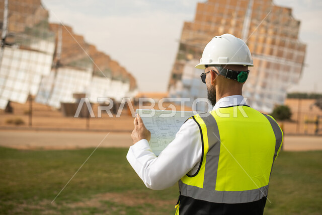 صورة مقربة لمهندس عربي خليجي سعودي يشرف على محطات الطاقة الشمسية في السعودية، استخدام جهاز الحاسب المحمول , إرتداء خوذة وسترة الحماية الخاصة بالعمل، مخططات وتصاميم هندسية، الواح وستلايت انتاج الطاقة الكهربائية , محطات الطاقة الشمسية المتجددة .