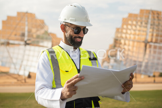 محطات الطاقة الشمسية المتجددة، صورة مقربة لمهندس عربي خليجي سعودي يشرف على محطات الطاقة الشمسية في السعودية،  إرتداء خوذة وسترة الحماية الخاصة بالعمل، مخططات وتصاميم هندسية، الواح وستلايت انتاج الطاقة الكهربائية .