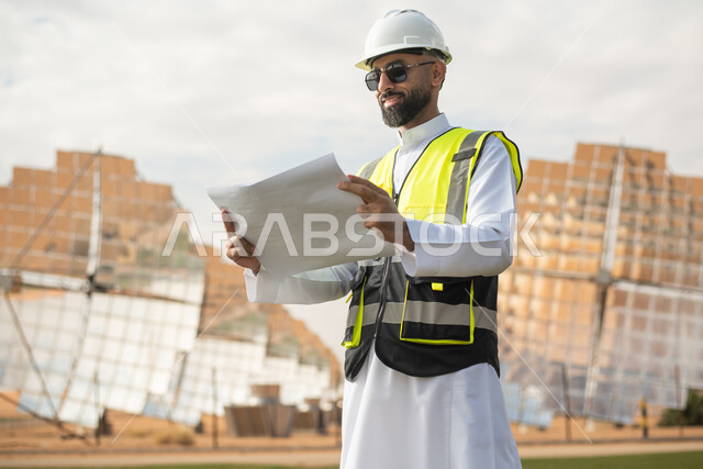 مهندس عربي خليجي سعودي يشرف على محطات الطاقة الشمسية في السعودية،  إرتداء خوذة وسترة الحماية الخاصة بالعمل، مخططات وتصاميم هندسية،  محطات الطاقة الشمسية المتجددة، الواح وستلايت انتاج الطاقة الكهربائية .