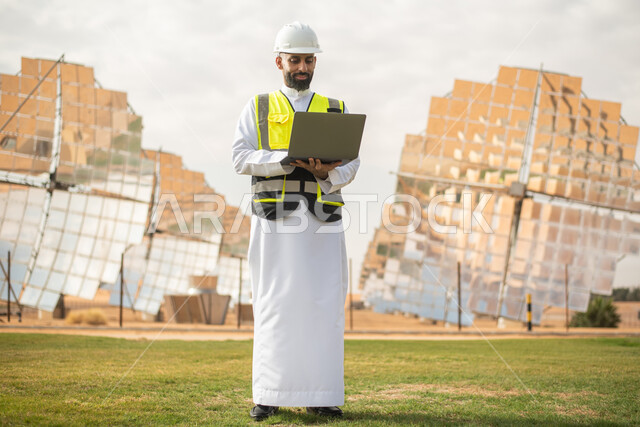 مهندس عربي خليجي سعودي يشرف على محطات الطاقة الشمسية في السعودية، استخدام جهاز الحاسب المحمول , إرتداء خوذة وسترة الحماية الخاصة بالعمل، مخططات وتصاميم هندسية،  محطات الطاقة الشمسية المتجددة، ,الواح وستلايت انتاج الطاقة الكهربائية . 