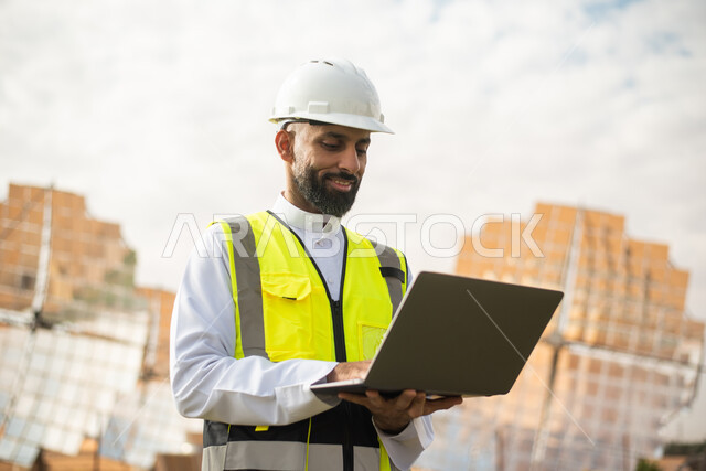 استخدام جهاز الحاسب المحمول , مهندس عربي خليجي سعودي يشرف على محطات الطاقة الشمسية في السعودية، إرتداء خوذة وسترة الحماية الخاصة بالعمل، مخططات وتصاميم هندسية،  محطات الطاقة الشمسية المتجددة، الواح وستلايت انتاج الطاقة الكهربائية . 