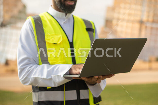 صورة مقربة لمهندس عربي خليجي سعودي يستخدم جهاز الحاسب المحمول , إرتداء خوذة وسترة الحماية الخاصة بالعمل، مخططات وتصاميم هندسية،  محطات الطاقة الشمسية المتجددة، الواح وستلايت انتاج الطاقة الكهربائية . 
