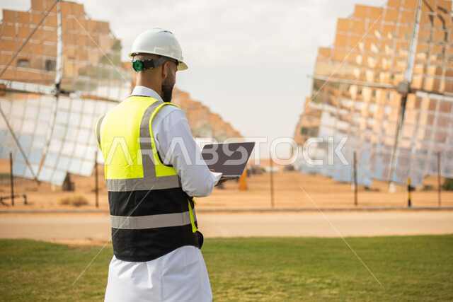 صورة من الخلف لمهندس عربي خليجي سعودي يشرف على محطات الطاقة الشمسية في السعودية، استخدام جهاز الحاسب المحمول , إرتداء خوذة وسترة الحماية الخاصة بالعمل، مخططات وتصاميم هندسية،  محطات الطاقة الشمسية المتجددة، الواح وستلايت انتاج الطاقة الكهربائية . 