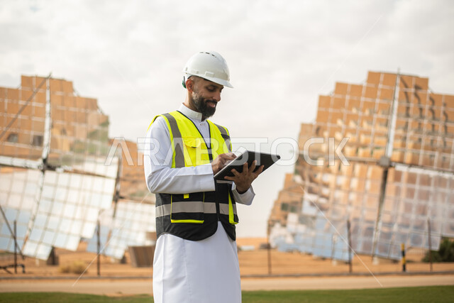 صورة مقربة لمهندس عربي خليجي سعودي يشرف على محطات الطاقة الشمسية في السعودية، استخدام جهاز التابلت في ادارة المشاريع , إرتداء خوذة وسترة الحماية الخاصة بالعمل، مخططات وتصاميم هندسية،  محطات الطاقة الشمسية المتجددة، الواح وستلايت انتاج الكهرباء . 