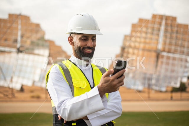 صورة مقربة لمهندس عربي خليجي سعودي يشرف على محطات الطاقة الشمسية في السعودية، استخدام الهاتف المحمول , إرتداء خوذة وسترة الحماية الخاصة بالعمل، استخدام التقنية والتكنولوجيا في المشاريع المستقبلية ،  محطات الطاقة الشمسية المتجددة، الواح وستلايت .