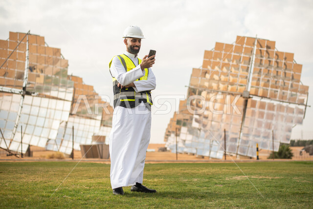مهندس عربي خليجي سعودي يشرف على محطات الطاقة الشمسية في السعودية، استخدام الهاتف المحمول , إرتداء خوذة وسترة الحماية الخاصة بالعمل، استخدام التقنية والتكنولوجيا في المشاريع المستقبلية ،  محطات الطاقة الشمسية المتجددة، الواح وستلايت انتاج الطاقة الكهربائية