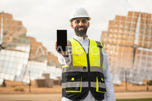 صورة مقربة لمهندس عربي خليجي سعودي يشرف على محطات الطاقة الشمسية في السعودية، استخدام التقنية والتكنولوجيا في المشاريع المستقبلية , إرتداء خوذة وسترة الحماية الخاصة بالعمل، استتخدام الهاتف المحمول ،عرض شاشة سوداء فارغة ،  محطات الطاقة الشمسية المتجددة .