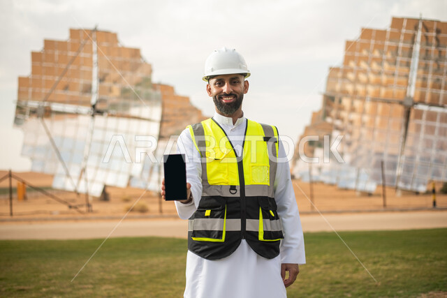 مهندس عربي خليجي سعودي يشرف على محطات الطاقة الشمسية في السعودية، استخدام التقنية والتكنولوجيا في المشاريع المستقبلية , إرتداء خوذة وسترة الحماية الخاصة بالعمل، استتخدام الهاتف المحمول ،عرض شاشة سوداء فارغة ،  محطات الطاقة الشمسية المتجددة، الواح وستلايت 