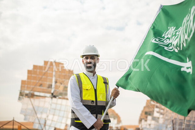 مهندس عربي خليجي سعودي يحمل علم المملكة العربية السعودية ، يوم العلم ,  محطات توليد الكهرباء عبر الطاقة الشمسية، الاحتفال باليوم الوطني السعودي 92، ستلايت والواح الطاقة الطبيعية المتجددة , رؤية ومشاريع مستقبلية جديدة .