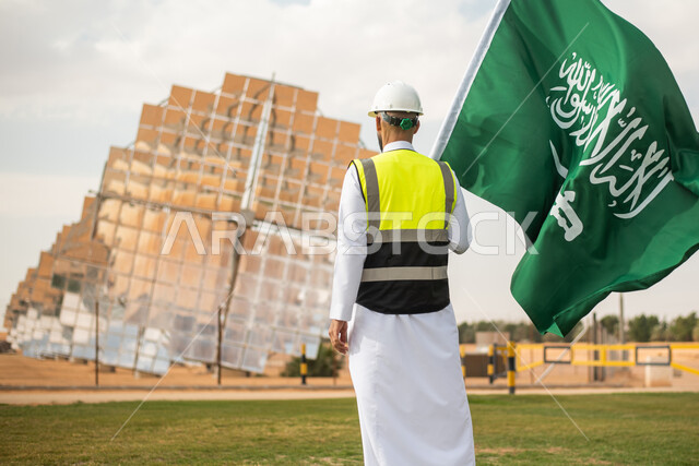 صورة من الخلف لمهندس عربي خليجي سعودي يحمل علم المملكة العربية السعودية ، يوم العلم ,  محطات توليد الكهرباء عبر الطاقة الشمسية، الاحتفال باليوم الوطني السعودي 92، ستلايت والواح الطاقة الطبيعية المتجددة , رؤية ومشاريع مستقبلية جديدة .