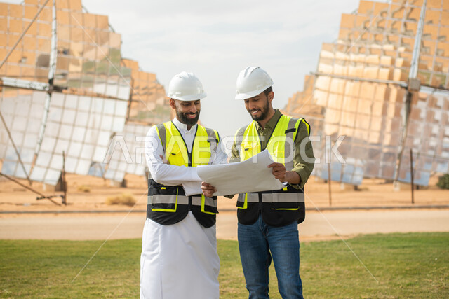 صورة مقربة لمهندسان عربيان خليجيان سعوديان يشرفان على محطات الطاقة الشمسية في السعودية، إرتداء خوذة وسترة الحماية الخاصة بالعمل، مخططات وتصاميم هندسية، حوارات ونقاشات عمل ،  محطات الطاقة الشمسية المتجددة، الواح وستلايت انتاج الطاقة الكهربائية .