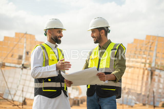 صورة مقربة لمهندسان عربيان خليجيان سعوديان يشرفان على محطات الطاقة الشمسية في السعودية، إرتداء خوذة وسترة الحماية الخاصة بالعمل، مخططات وتصاميم هندسية، حوارات ونقاشات عمل ،  محطات الطاقة الشمسية المتجددة، الواح وستلايت انتاج الطاقة الكهربائية .