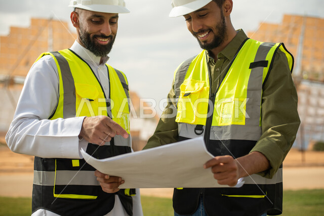 مهندسان عربيان خليجيان سعوديان يشرفان على محطات الطاقة الشمسية في السعودية، إرتداء خوذة وسترة الحماية الخاصة بالعمل، مخططات وتصاميم هندسية، حوارات ونقاشات عمل ،  محطات الطاقة الشمسية المتجددة، الواح وستلايت انتاج الطاقة الكهربائية ,إيماءات وجه مبتسمة .