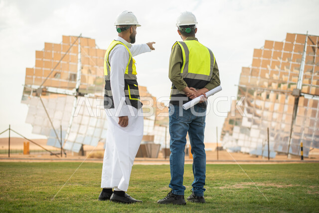 صورة من الخلف لمهندسان عربيان خليجيان سعوديان يشرفان على محطات الطاقة الشمسية في السعودية، إرتداء خوذة وسترة الحماية الخاصة بالعمل، مخططات وتصاميم هندسية، حوارات ونقاشات عمل ،  محطات الطاقة الشمسية المتجددة، الواح وستلايت انتاج الطاقة الكهربائية.
