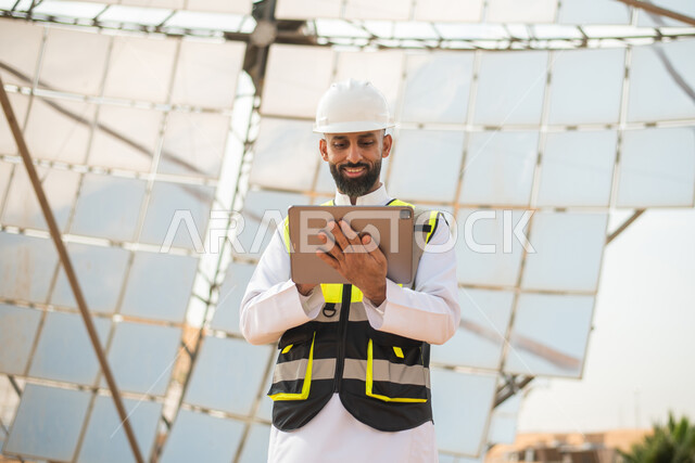 صورة مقربة من الاسفل لمهندس عربي خليجي سعودي يشرف على محطات الطاقة الشمسية في السعودية، استخدام جهاز التابلت في ادارة المشاريع , إرتداء خوذة وسترة الحماية الخاصة بالعمل، مخططات وتصاميم هندسية،محطات الطاقة الشمسية المتجددة، الواح وستلايت انتاج الكهرباء