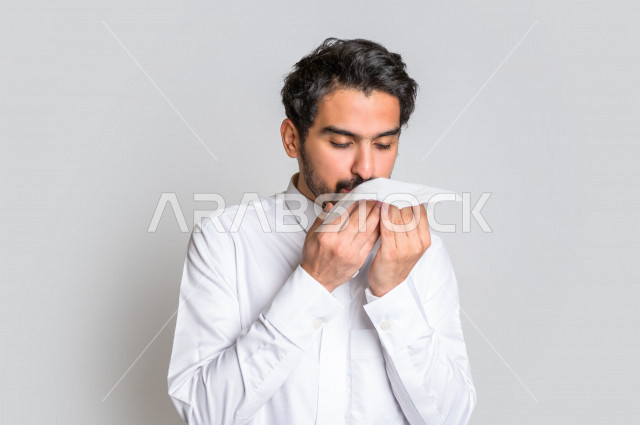 بورتريه لرجل عربي سعودي خليجي يرتدي الثوب السعودي , يحمل بيده مناديل ورقيه , إيماءات وجه تعبر عن المرض ، اشارات يدين تدل على العطاس والتعب , الإنفلونزا , فايروس كورونا , كوفيد 19 , خلفية بيضاء